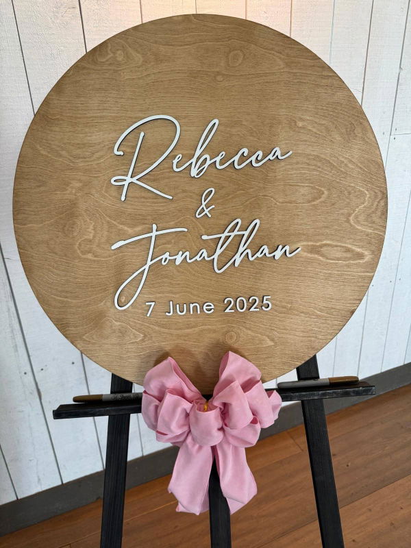 Customer photo: a round wooden guest book sign with two first names and a wedding date on an easel with a pink bow