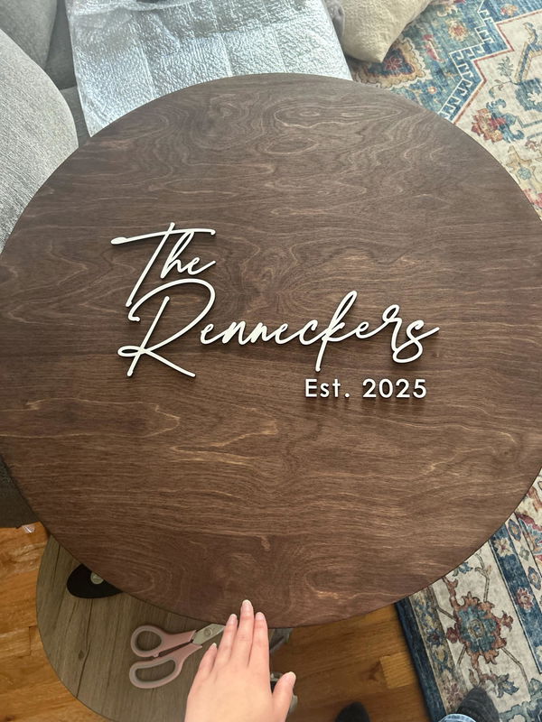 Customer photo: a dark round wooden family name sign with raised white lettering, held up at home