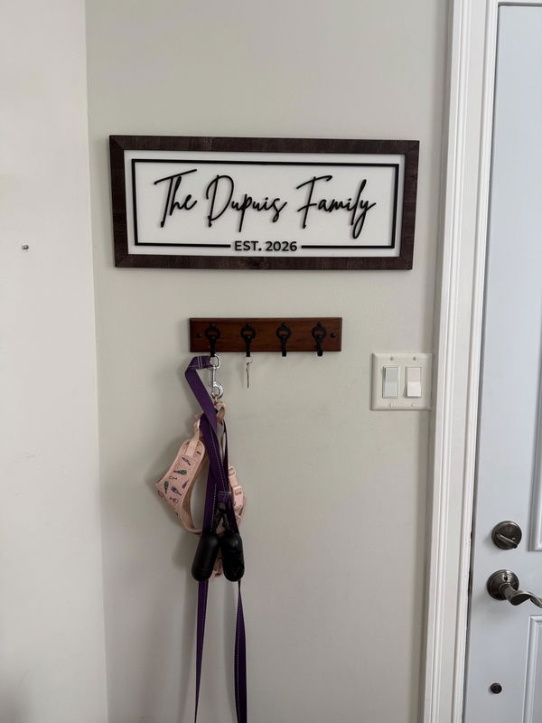 Customer photo: a framed family name sign reading The Dupuis Family, Est. 2026, on an entryway wall above a row of coat hooks.