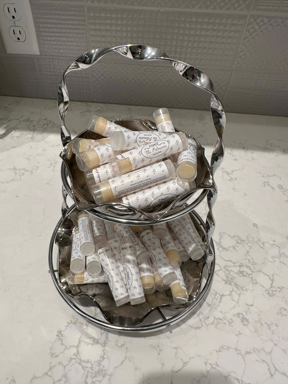 Dozens of custom-labeled lip balm favors piled on a two-tier silver stand, ready to set out for guests