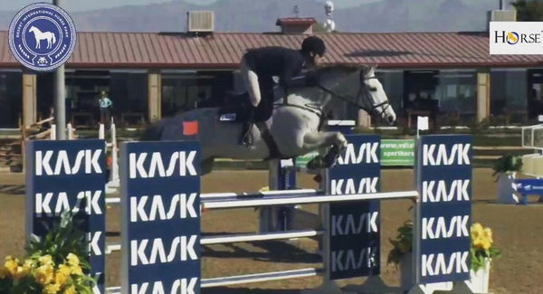 Rider on a gray horse jumping at a show with KASK banners